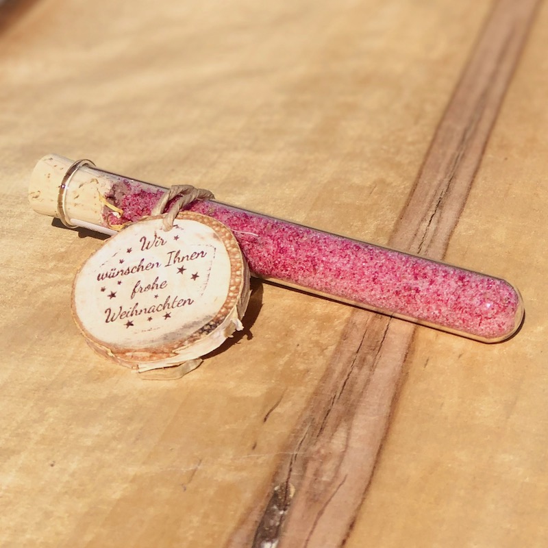 Glasröhrchen mit rotem Blütensalz und Holzanhänger mit der Beschriftung: Wir wünschen Ihnen frohe Weihnachten, als Giveaway .Auf einem Tisch liegend mit Lichterkette und Strohstern