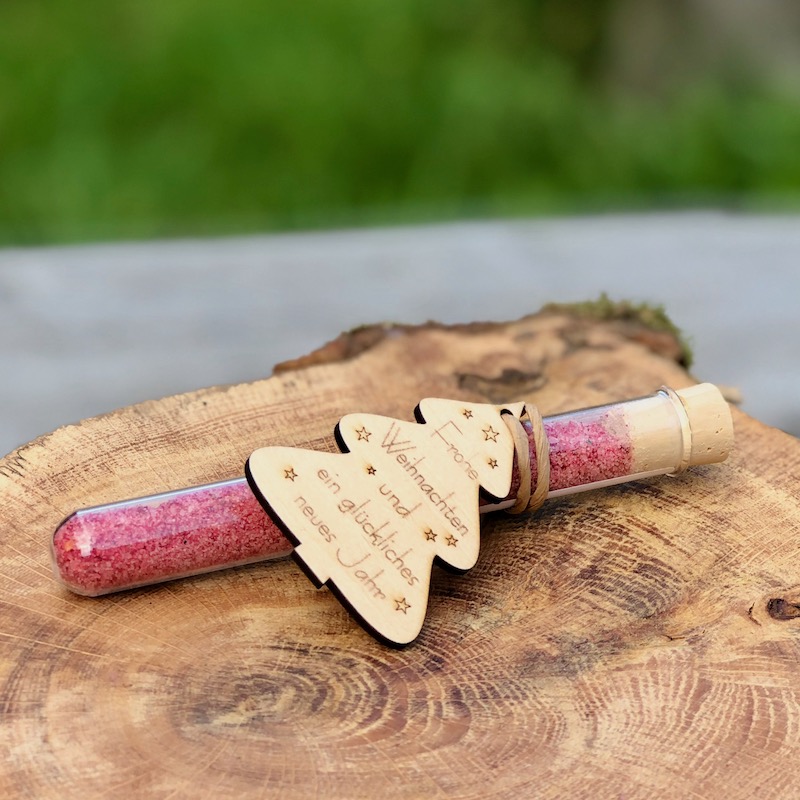 Glasröhrchen mit Blütensalz und Holzanhänger mit dem Aufdruck "frohe Weihnachten und ein glückliches neues Jahr" auf einem Tisch liegend mit Lichterkette und Strohstern